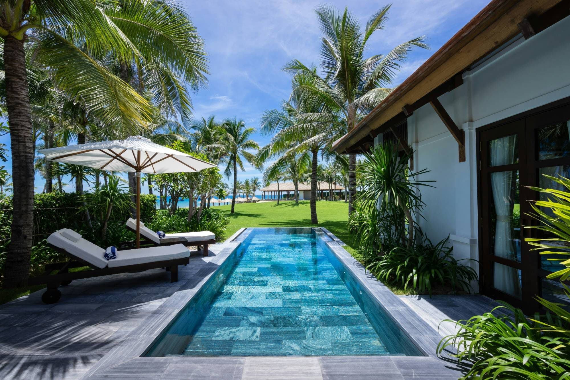 Private lap pool with sun loungers and a parasol in a palm-shaded villa garden near the sea